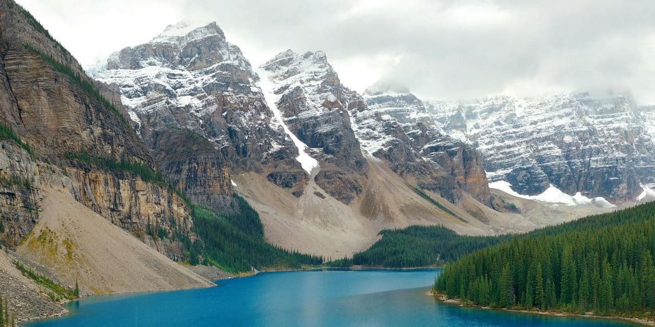 Un lac alpin d'un bleu éclatant se trouve au pied de montagnes imposantes et enneigées, avec une forêt dense de conifères le long de ses rives.