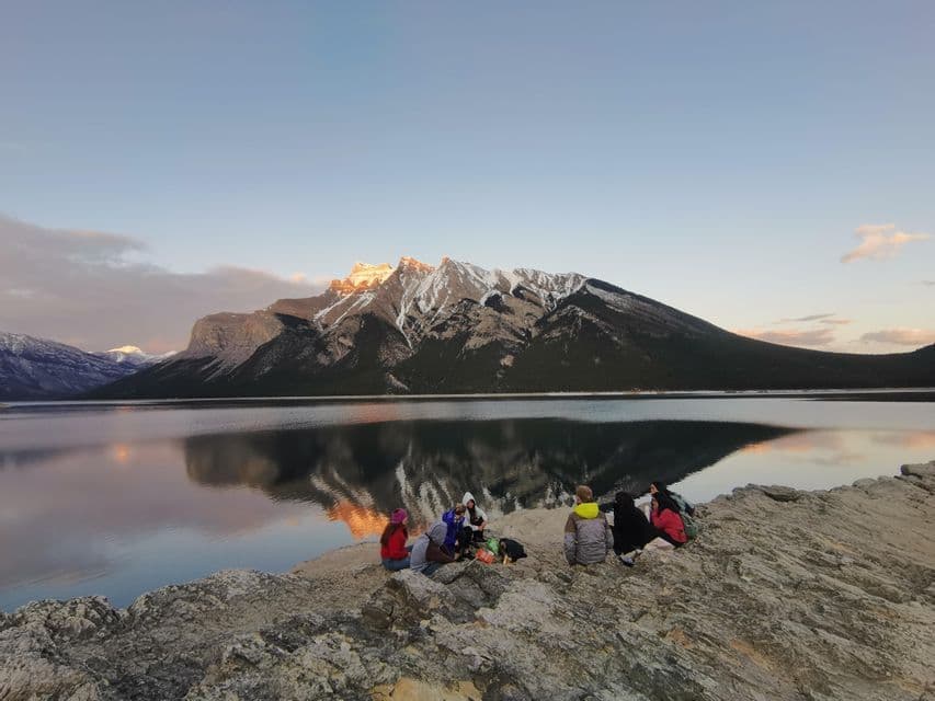 Un groupe WeRoad sur une rive rocheuse, au bord d'un lac calme, face à des montagnes enneigées au coucher du soleil.