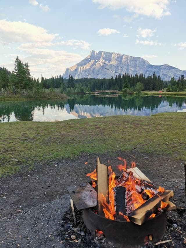 Un feu de camp brûle dans un foyer au bord d'un lac, avec une montagne et une forêt se reflétant dans l'eau calme sous un ciel partiellement nuageux.