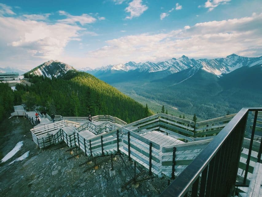 Une passerelle en bois sur le flanc d'une montagne surplombant une vallée avec des forêts de pins et des sommets enneigés lointains sous un ciel partiellement nuageux.