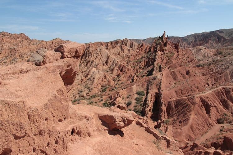Un paysage de formations rocheuses rouges déchiquetées et de canyons avec une végétation verte éparse sous un ciel bleu.