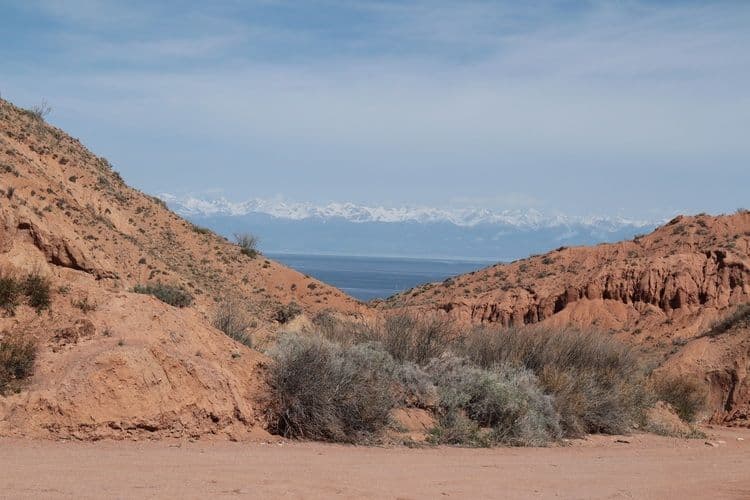 Une vue entre des collines de terre rouge montre une étendue d'eau lointaine et une chaîne de montagnes enneigée sous un ciel bleu.