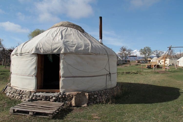 Une yourte blanche avec une fondation en pierre se dresse dans un campement avec des champs herbeux et des montagnes enneigées au loin.