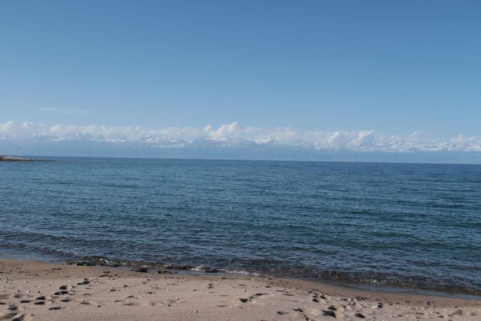 Une plage de sable avec une mer bleue calme face à des montagnes enneigées lointaines sous un ciel clair.