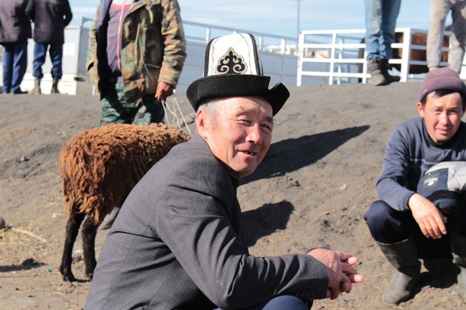 Un homme portant un chapeau brodé traditionnel sourit à un marché aux bestiaux à côté d'un mouton brun.