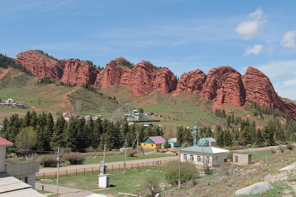 Un petit village avec une mosquée se trouve au pied d'une grande chaîne de montagnes rocheuses rouges et stratifiées sous un ciel bleu.