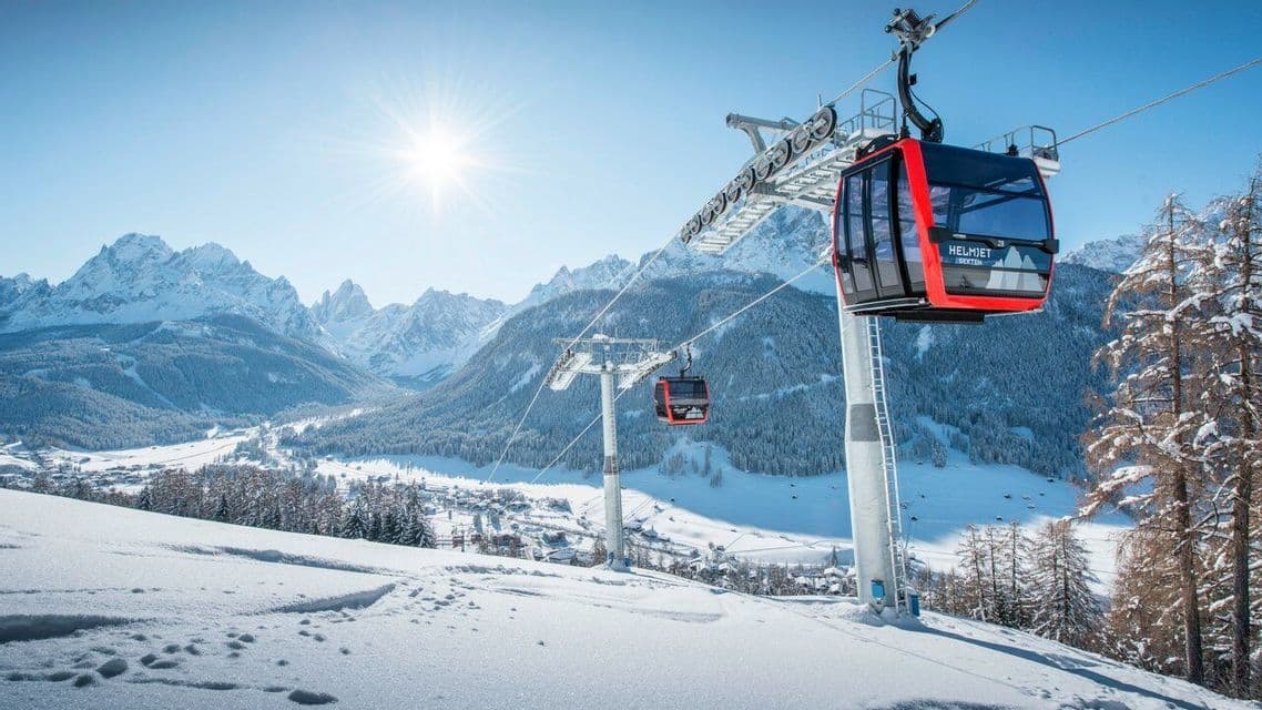 Gondolas rojas ascienden por una montaña nevada bajo un sol brillante, con una vasta cordillera nevada al fondo.