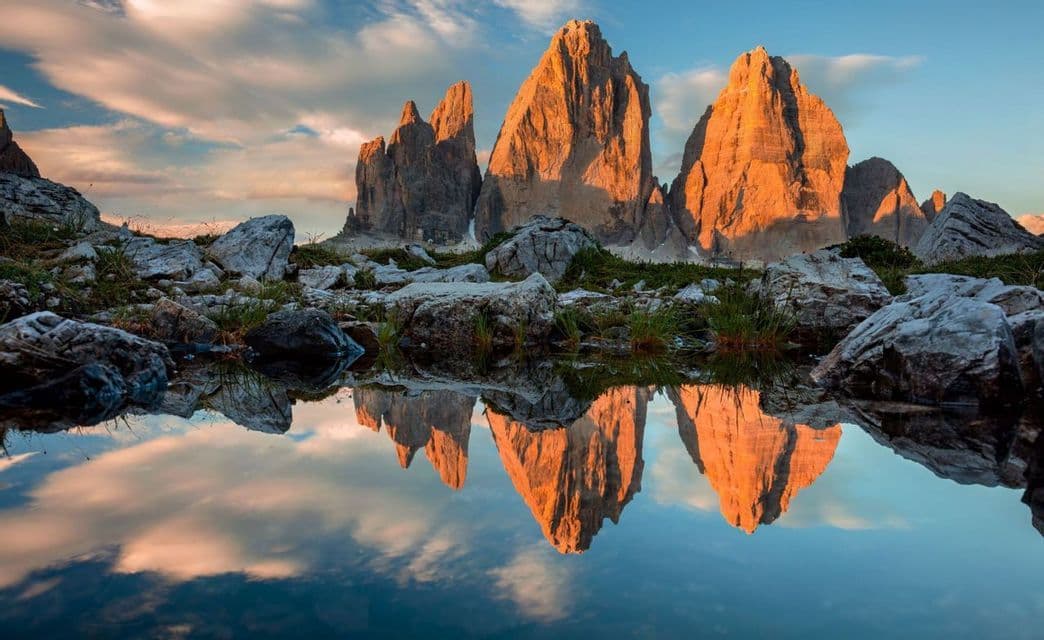 Picos montañosos irregulares iluminados por luz dorada se reflejan en un lago tranquilo con una orilla rocosa en primer plano.