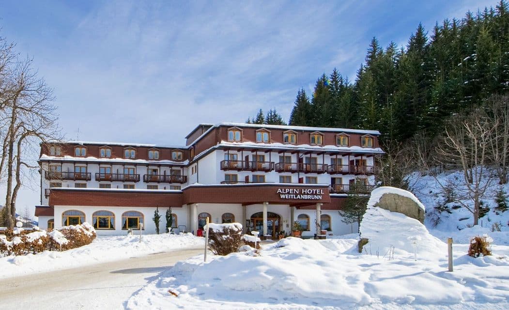 Un gran hotel alpino de varios pisos se alza en un paisaje nevado al pie de un bosque de pinos bajo un cielo parcialmente nublado.
