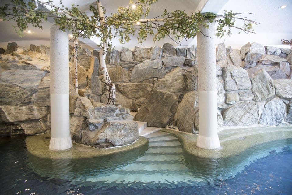 Peldaños de azulejos conducen a una piscina cubierta rodeada por un gran muro de roca, columnas blancas y abedules decorativos.