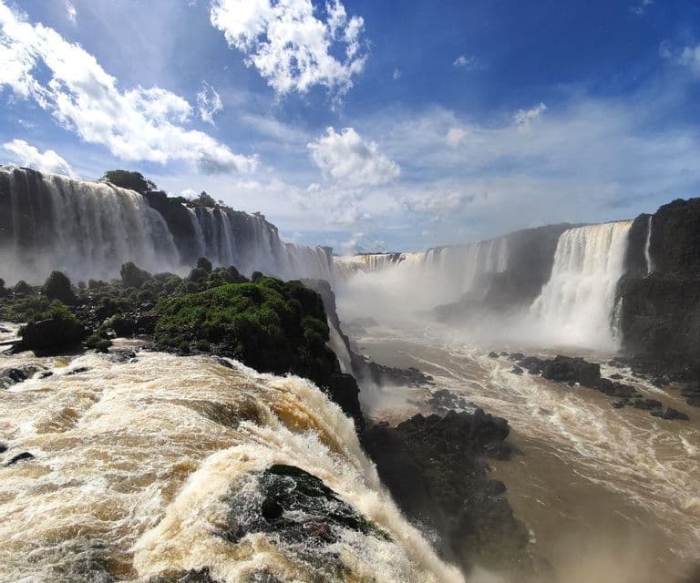 Ein weitläufiger Blick auf ein großes Wasserfallsystem, das in eine neblige Schlucht stürzt, mit Stromschnellen im Vordergrund unter einem teilweise bewölkten Himmel.