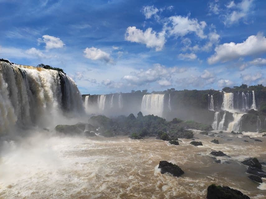 Potenti cascate si gettano in un fiume marrone turbolento, con nebbia che sale contro una fitta vegetazione verde, sotto un cielo blu e nuvoloso.