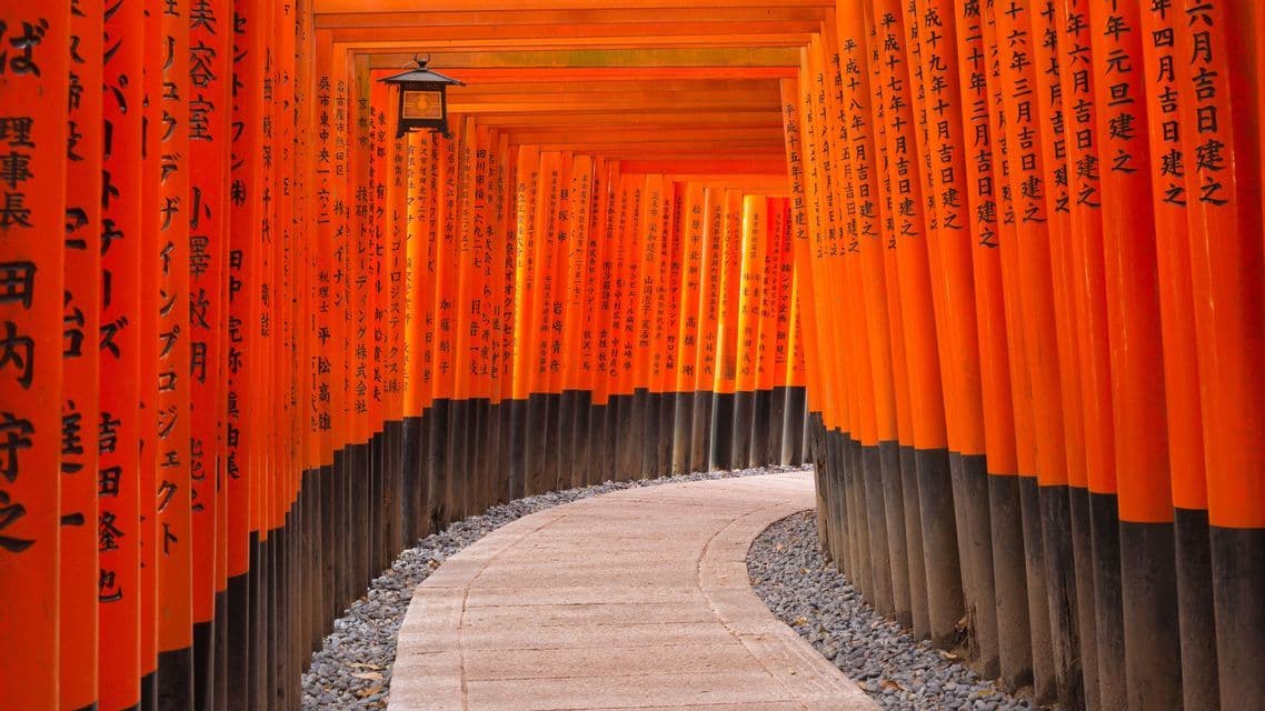A curving stone path leads through a corridor of bright orange Torii gates inscribed with Japanese calligraphy.