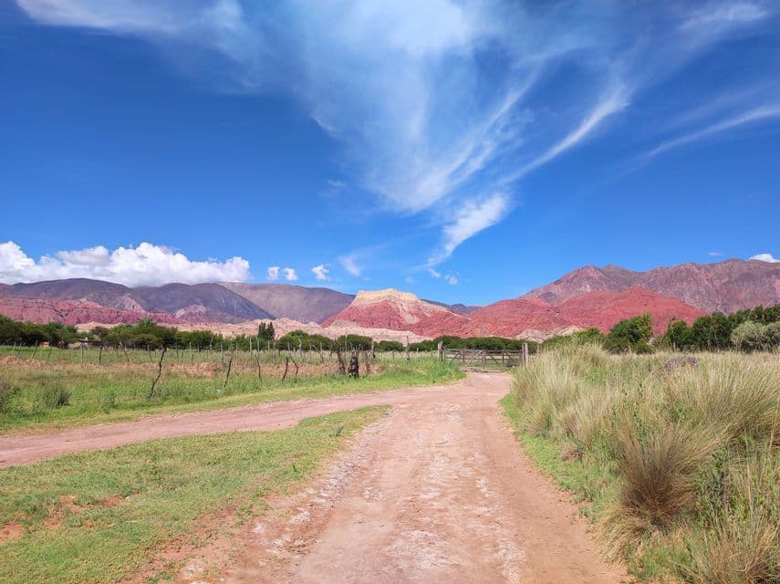 Una strada sterrata attraversa un campo erboso verso una catena di montagne rosse e gialle sotto un cielo azzurro con nuvole leggere.
