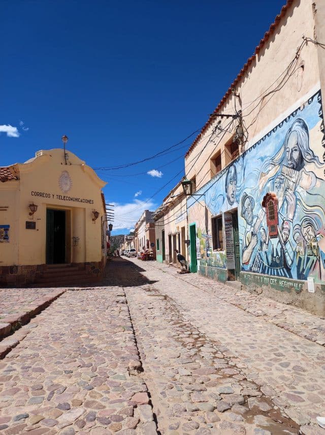Una calle adoquinada pasa entre un edificio amarillo y otro con un gran mural colorido bajo un cielo azul claro.