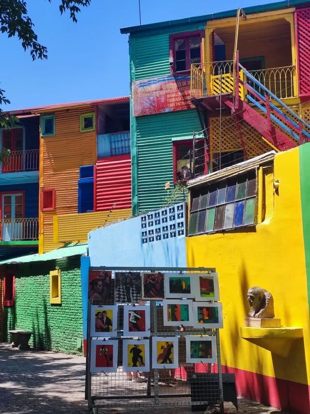 Una scena di strada illuminata dal sole con edifici dai colori vivaci in lamiera ondulata e una rastrelliera di stampe d'arte di tango in vendita.