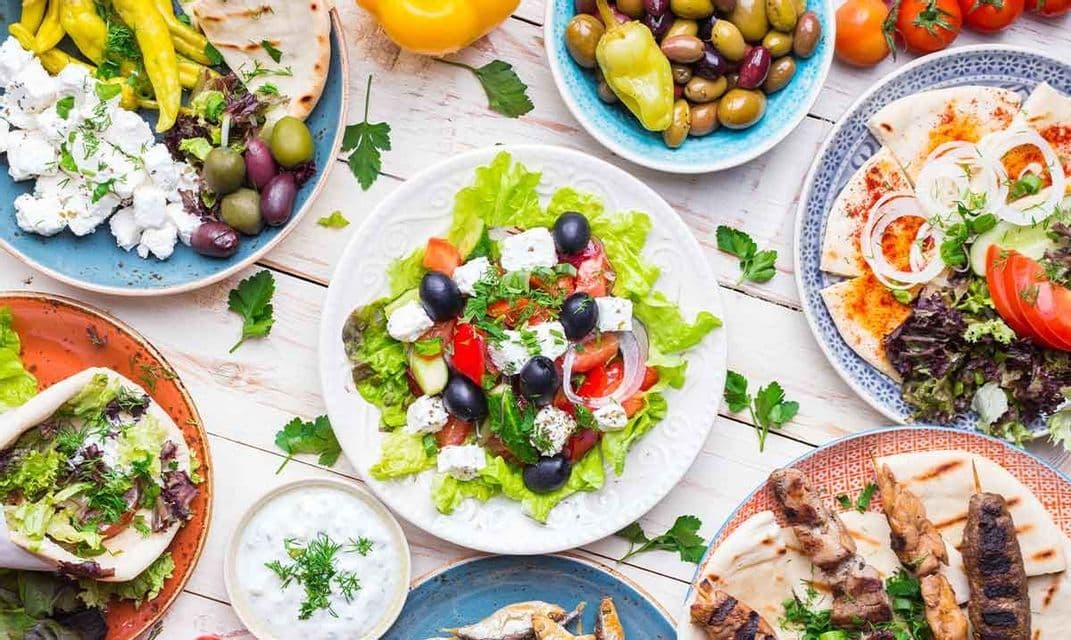 Eine Draufsicht auf einen Tisch mit griechischen Gerichten, darunter Feta-Salat, Souvlaki, Pitabrot und Oliven.