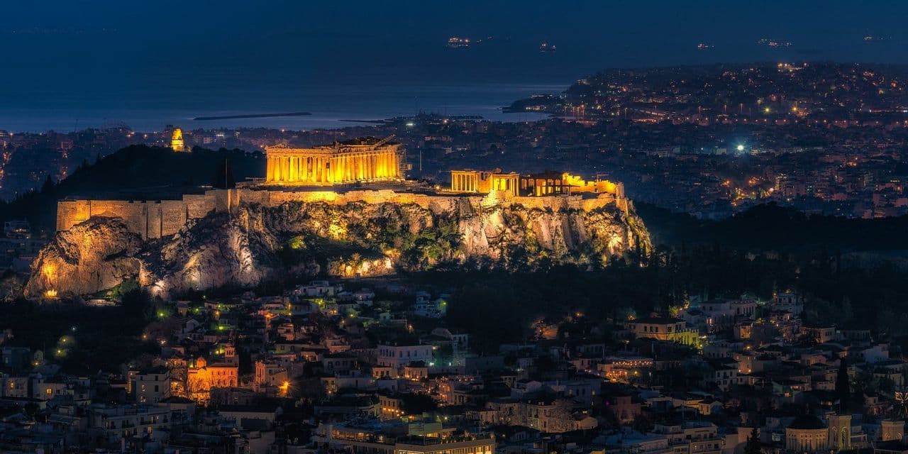 Die antike Akropolis erstrahlt nachts hell, überblickt die weitläufigen Lichter der Stadt und das Meer in der Ferne.