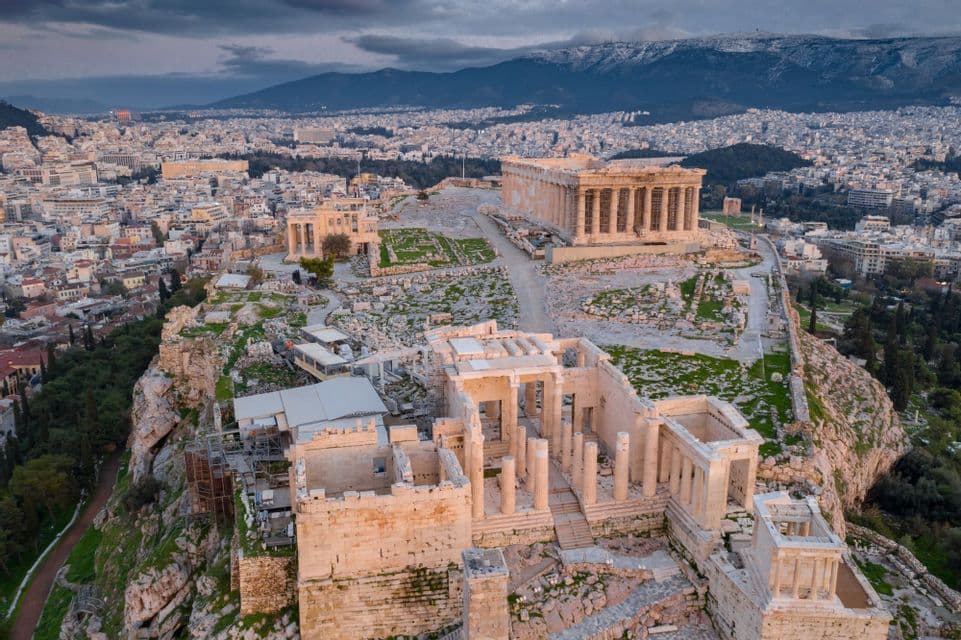 An aerial view of ancient ruins on a rocky citadel, overlooking a dense city with distant mountains under a cloudy sky.