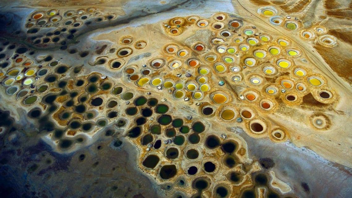 An aerial view of a landscape filled with numerous circular pools of water in shades of green, yellow, and orange.
