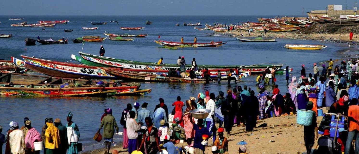 A crowd of people gathers on a sandy beach next to many colorful, traditional boats floating in the sea.