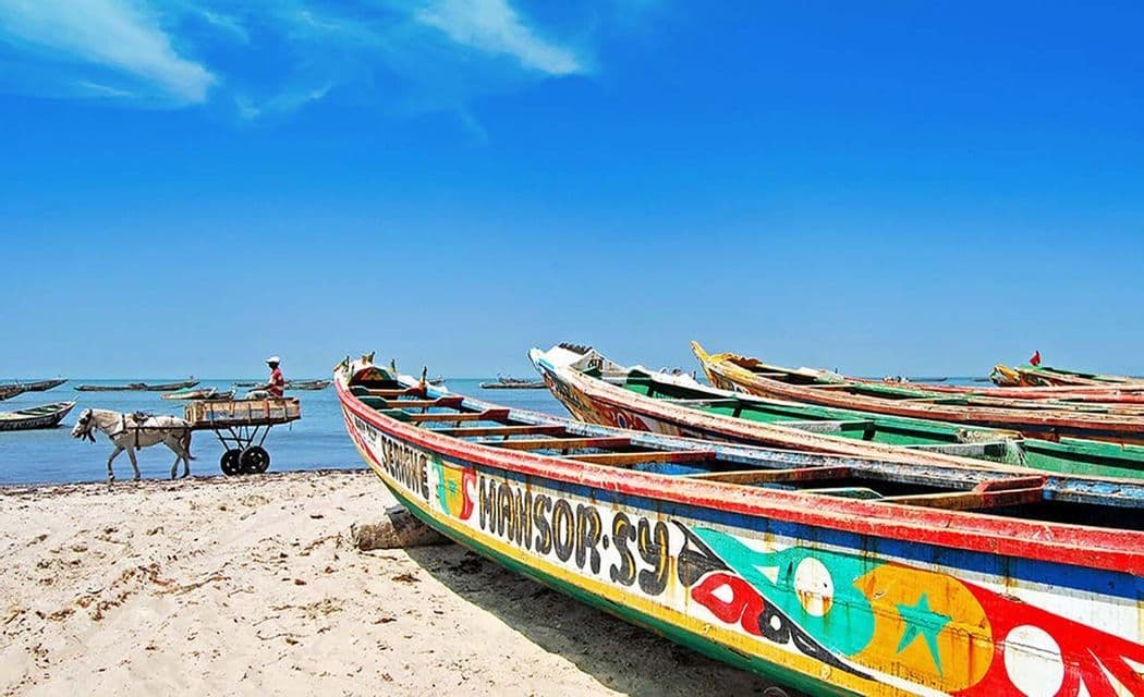 Colorful painted boats sit on a sandy beach as a horse-drawn cart passes by the water under a clear blue sky.