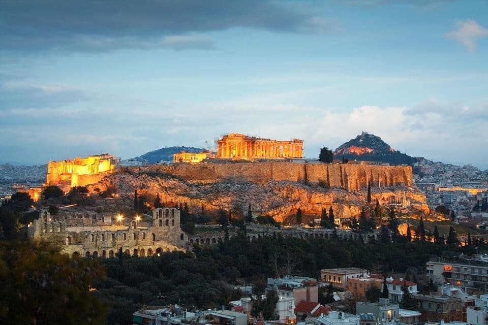 Una antigua ciudadela, brillantemente iluminada con luces doradas, se asienta sobre una colina rocosa con vistas a un paisaje urbano al anochecer.