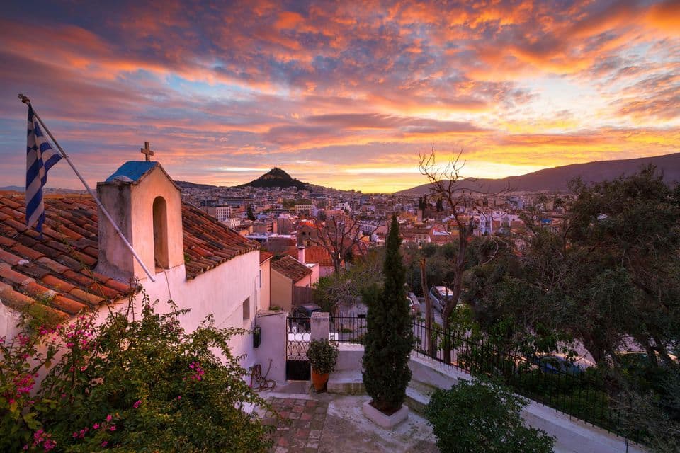 Una iglesia encalada con una bandera griega en su tejado domina un paisaje urbano con una colina lejana bajo un cielo de atardecer colorido.