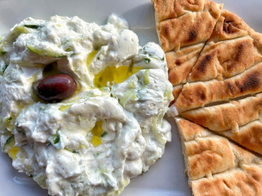 Tzatziki cremoso rociado con aceite de oliva y adornado con una aceituna, servido junto a pan de pita a la parrilla.