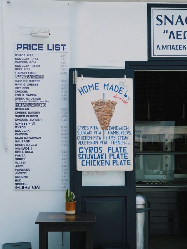 El escaparate de una tienda de gyros griega con una lista de precios en la pared y un cartel de menú pintado a mano.