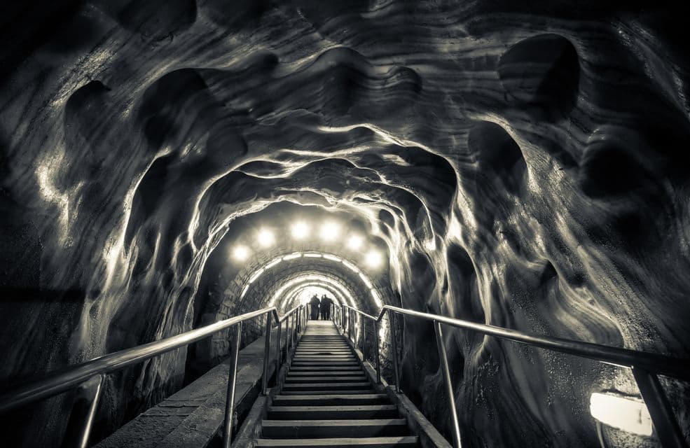 Un viaggio di gruppo WeRoad si trova in cima a una lunga scalinata all'interno di un tunnel illuminato con pareti di roccia scolpite, fotografato in bianco e nero.