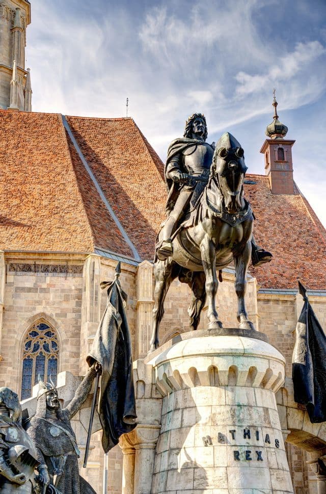 Inquadratura dal basso della statua equestre in bronzo di Re Mattia davanti a una chiesa con tetto in tegole rosse.