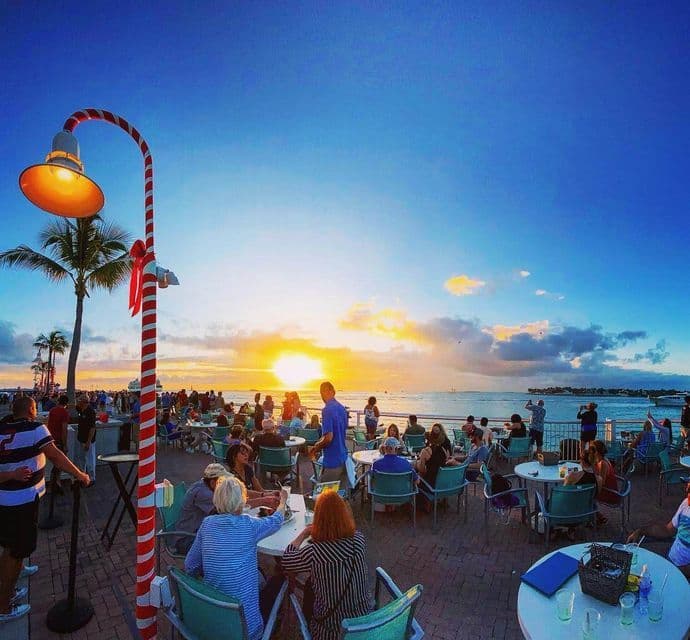 Una folla di persone si raduna su una terrazza lungomare al tramonto, con un lampione decorativo a strisce bianche e rosse in primo piano.