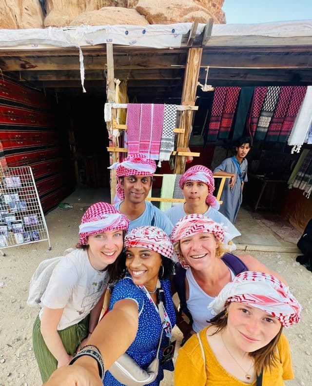 Un groupe WeRoad en voyage, portant des keffiehs assortis rouges et blancs, prend un selfie devant un stand de marché en plein air.