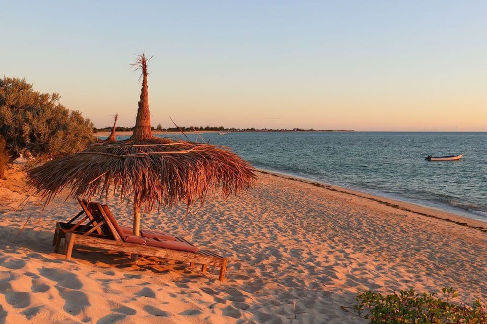 Ein Strohschirm und zwei Liegestühle an einem Sandstrand mit Meerblick bei goldenem Sonnenuntergang.