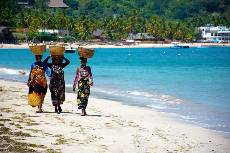 Three women in colorful clothing walk along a sandy beach, each carrying a woven basket on her head by a turquoise sea.