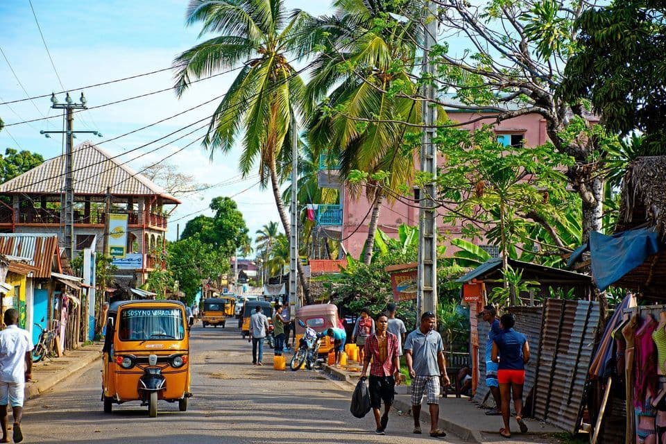 A bustling street scene with people walking, orange tuk-tuks, and buildings shaded by tall palm trees.