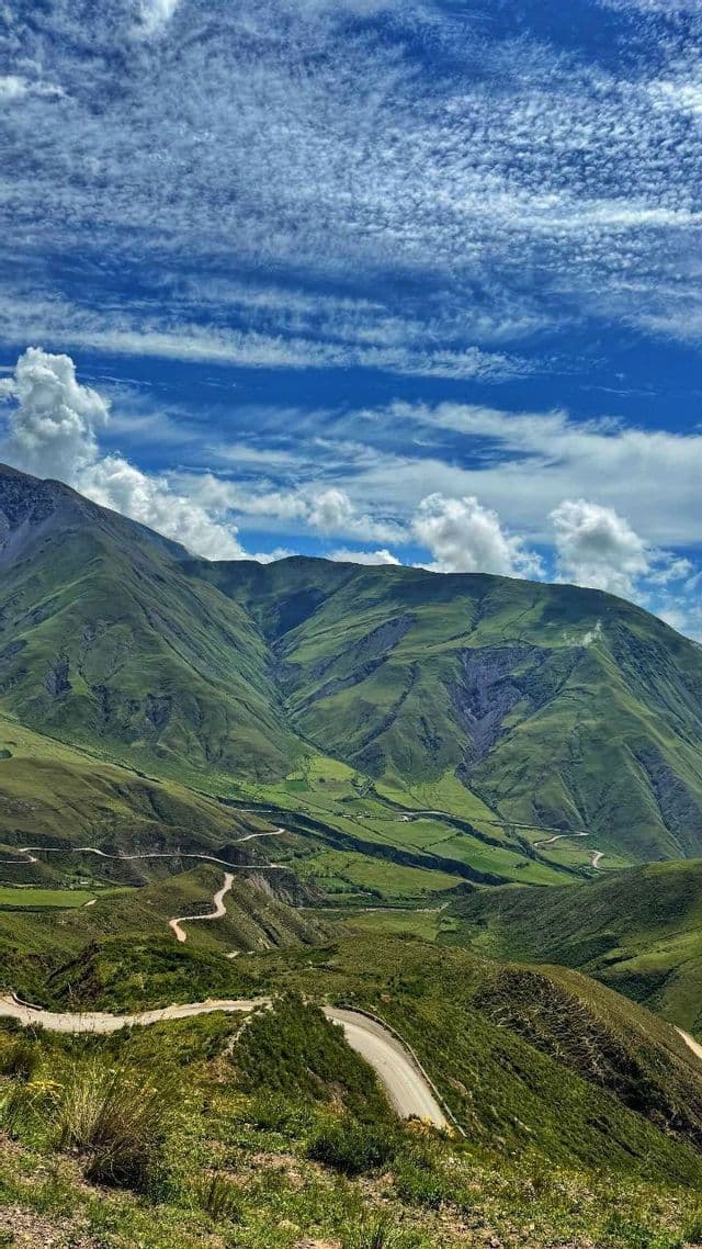 Una strada sterrata tortuosa si snoda attraverso montagne verdi e lussureggianti sotto un cielo blu con nuvole bianche sparse.