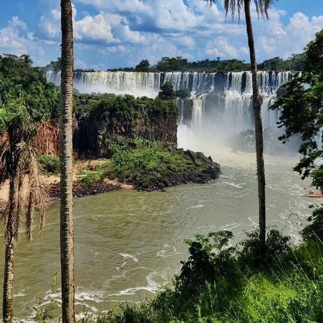 Un'ampia cascata si getta in un fiume, con una rigogliosa vegetazione verde su uno sperone roccioso e palme in primo piano.