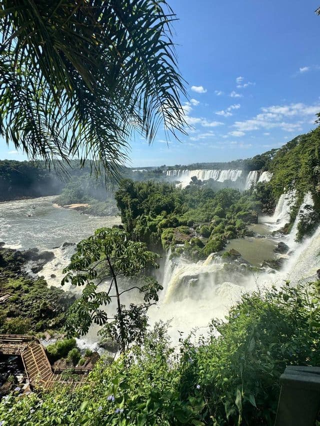 Vista panoramica di un grande sistema di cascate in una giungla lussureggiante, con l'acqua che si getta in un fiume sotto un cielo azzurro.