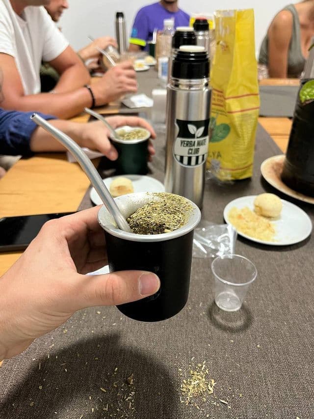 Primo piano di una mano che sorregge una tazza tradizionale di yerba mate, mentre un gruppo WeRoad condivide la bevanda attorno a un tavolo.