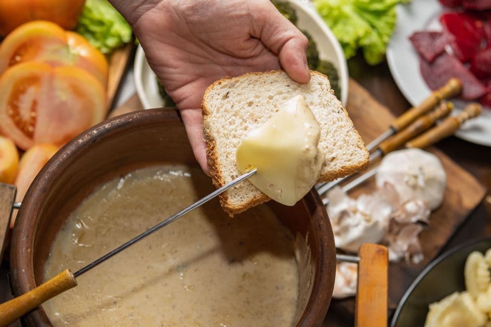 La mano di una persona intinge una patata su una forchetta da fonduta in una pentola di fonduta al formaggio, accanto a pane e verdure.