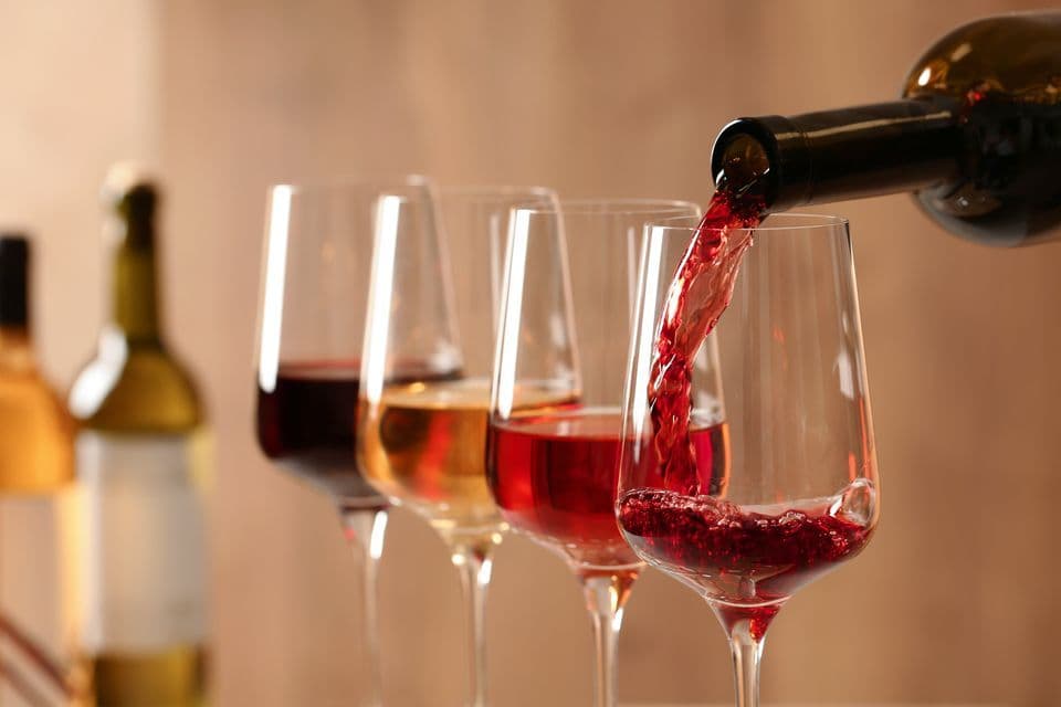 Primo piano di vino rosso versato da una bottiglia in un calice, con altri calici di vino allineati per una degustazione.