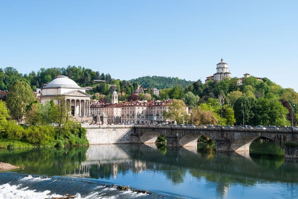 Vista di una città oltre il fiume, con un ponte ad arco in pietra, una chiesa a cupola e un edificio su una lussureggiante collina verde.