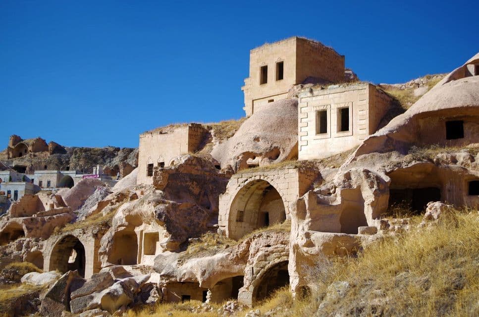 Des bâtiments anciens en grès et des habitations troglodytes sont sculptés dans une colline rocheuse sous un ciel bleu clair.