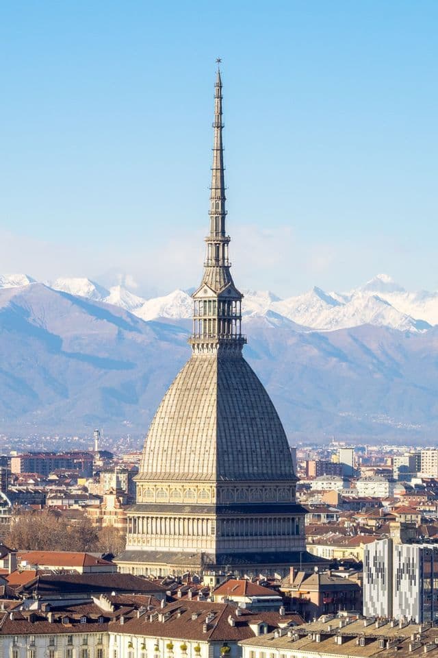 Un alto edificio a cupola con una guglia prominente si staglia su un paesaggio urbano, con montagne innevate visibili sullo sfondo.