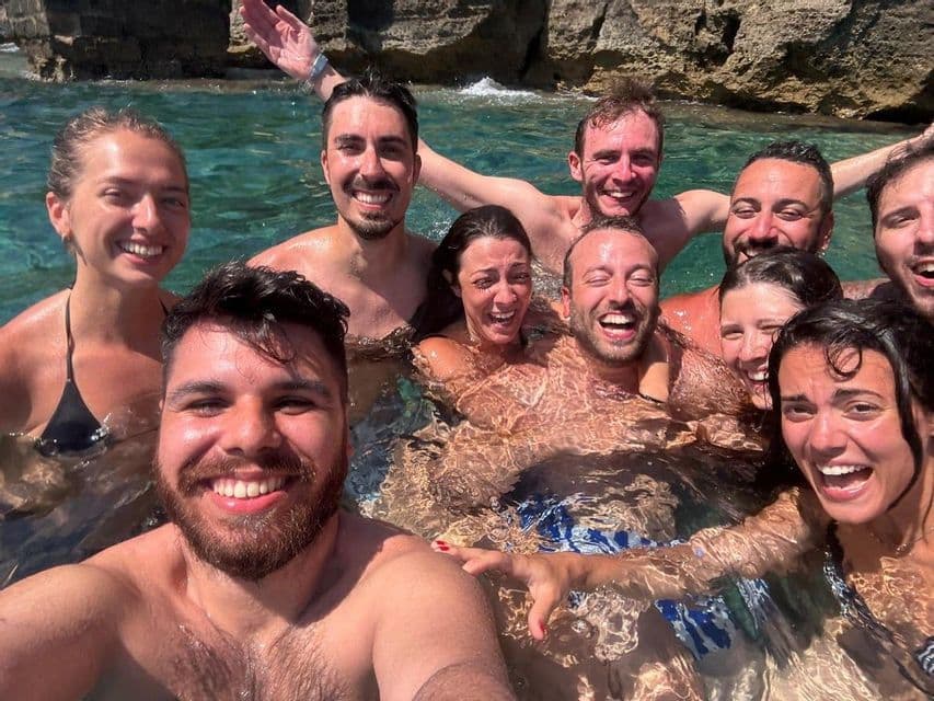 A smiling WeRoad group trip takes a selfie while swimming together in the clear sea near a rocky cliff.