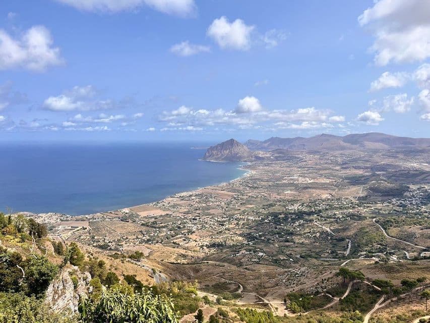 A high-angle view of a mountainous coastline, a town, and the blue sea under a partly cloudy sky.