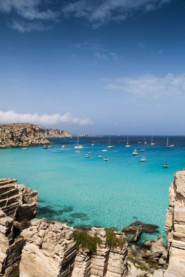 A high-angle view of numerous boats and sailboats anchored in a turquoise bay surrounded by rocky cliffs.
