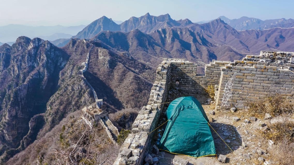 Une tente de camping verte installée sur un long mur de pierre en ruine serpentant à travers une vaste chaîne de montagnes escarpée sous un ciel clair.
